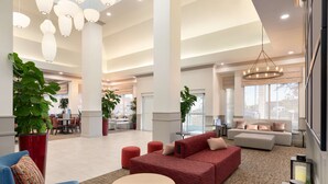 A spacious lobby with a red sofa, blue armchair, and a round table. There are large potted plants, a chandelier, and a view of the outside through large windows.
