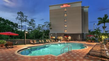 Zona de piscina con sillas de descanso y sombrillas, junto a un edificio Hilton Garden Inn.