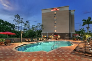 A swimming pool area with lounge chairs and umbrellas, surrounded by a Hilton Garden Inn building.