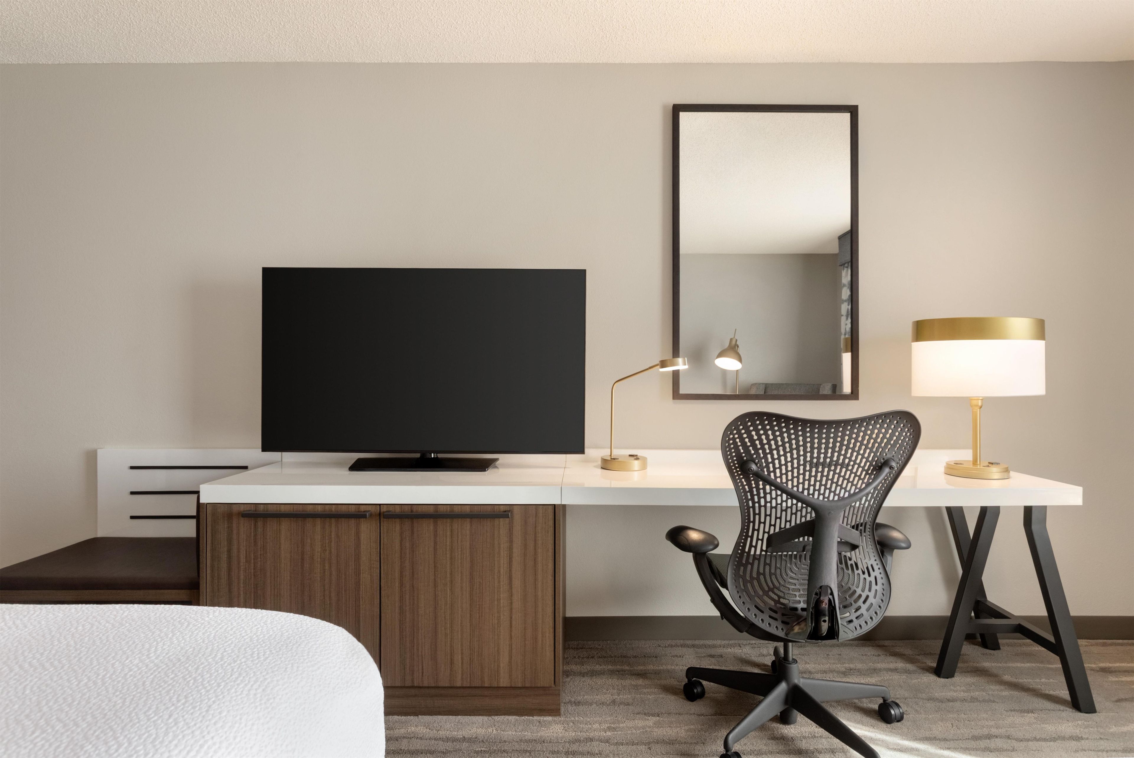 A hotel room with a flat-screen TV, a desk with a lamp, a chair, and a bed.