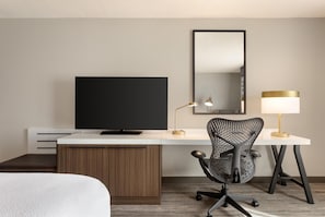 A hotel room with a flat-screen TV, a desk with a lamp, a chair, and a bed.
