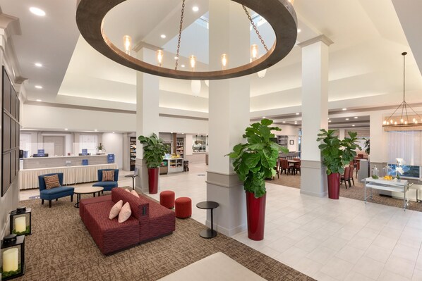 Un vestíbulo espacioso con diseño moderno, que presenta una zona de estar central con un sofá rojo y varios otomanes rojos, rodeado de grandes macetas con plantas y un candelabro circular.