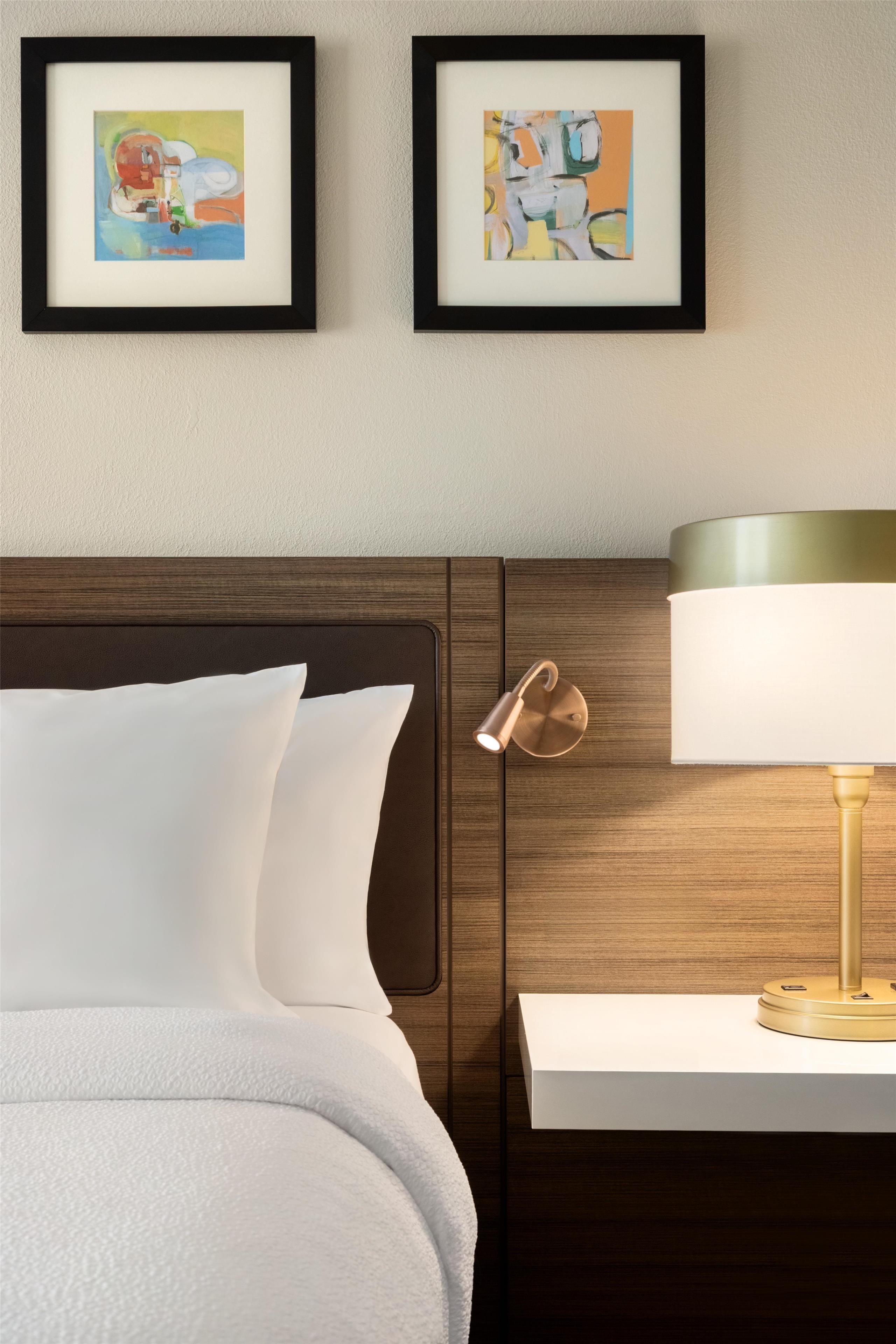 A neatly made bed with white linens, a wooden headboard, and a bedside table with a lamp. Two framed artworks are hung on the wall above the bed.