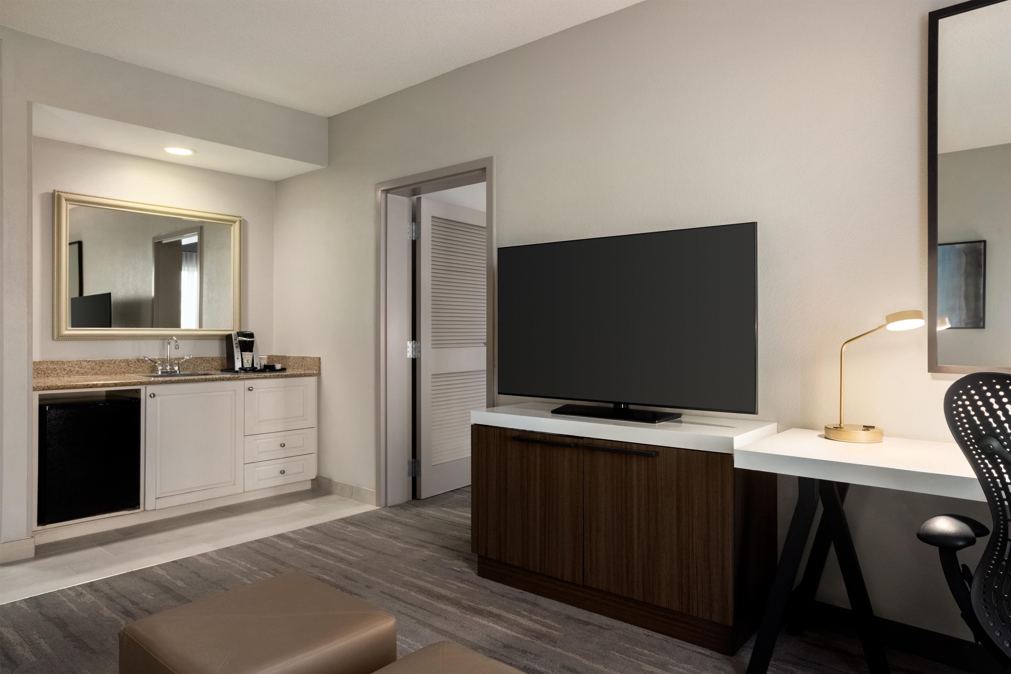 A modern hotel room with a kitchenette, a flat-screen TV, a desk with a lamp, and a chair.