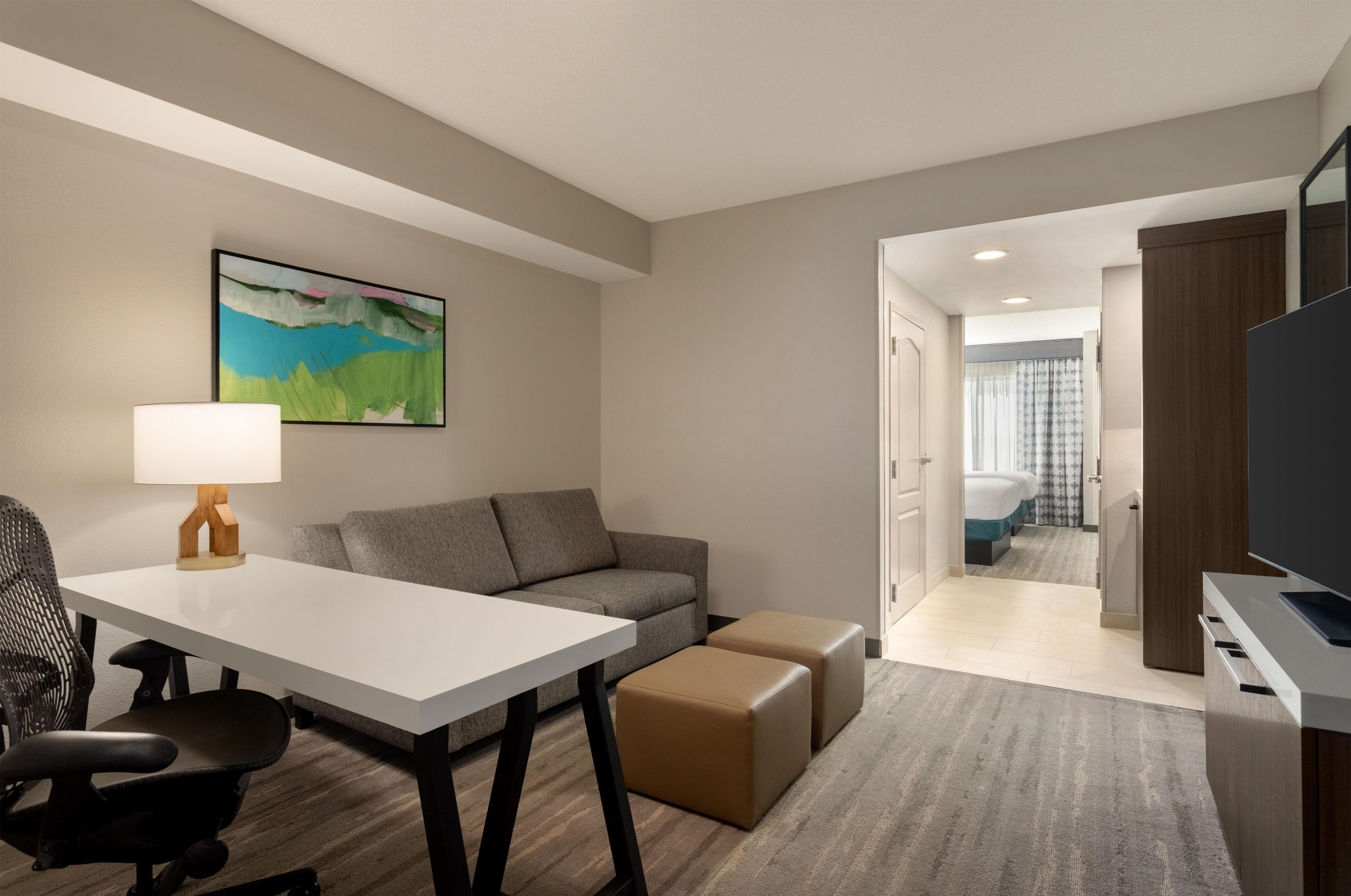 A hotel room with a white desk, a grey sofa, a brown ottoman, a black office chair, a painting on the wall, and a television on a stand.