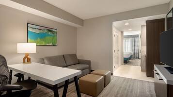 A hotel room with a white desk, a grey sofa, a brown ottoman, a black office chair, a painting on the wall, and a television on a stand.