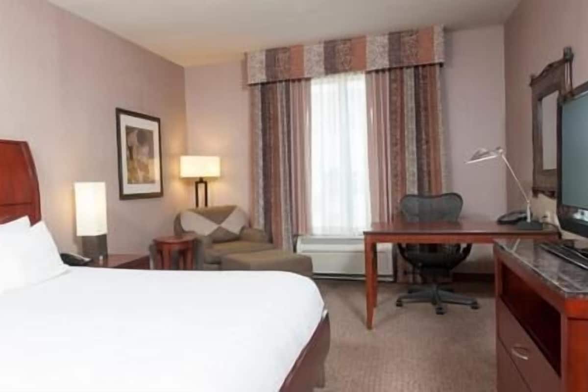 Ein Hotelzimmer mit Bett, Schreibtisch, Stuhl, Lampe, Fernseher und Vorhängen.