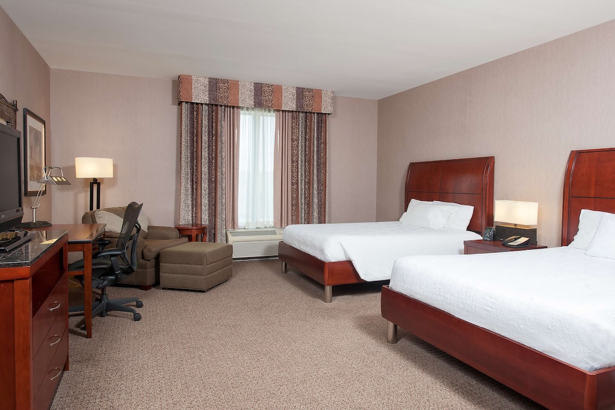 Ein Hotelzimmer mit zwei Betten, einem Schreibtisch, einem Stuhl, einem Fernseher und einem Fenster mit Vorhängen.
