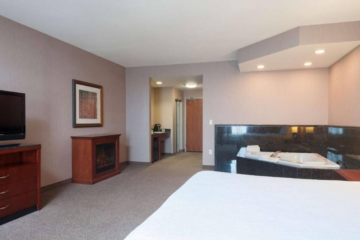 Ein Hotelzimmer mit Bett, Fernseher, Kamin, Badezimmer mit schwarzem Waschtisch und einem Gang zu einem weiteren Zimmer.