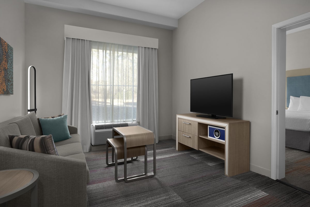A hotel room with a sofa, a small table, a TV on a wooden stand, and a window with curtains.