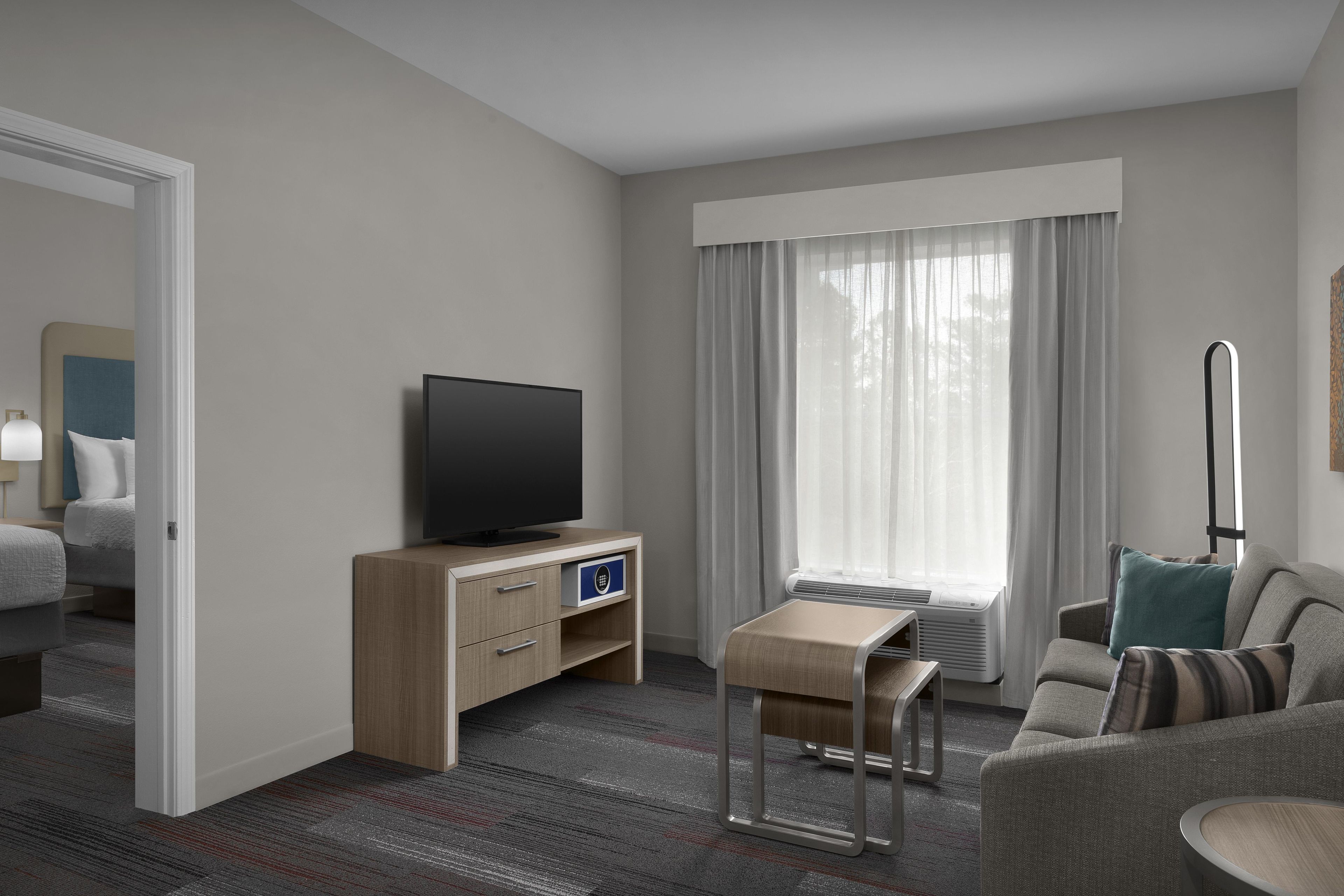 Ein Hotelzimmer mit Fernseher auf einer Holzkommode, einem kleinen Tisch mit einer Bank, einem Sofa und einem großen Fenster mit Vorhängen.