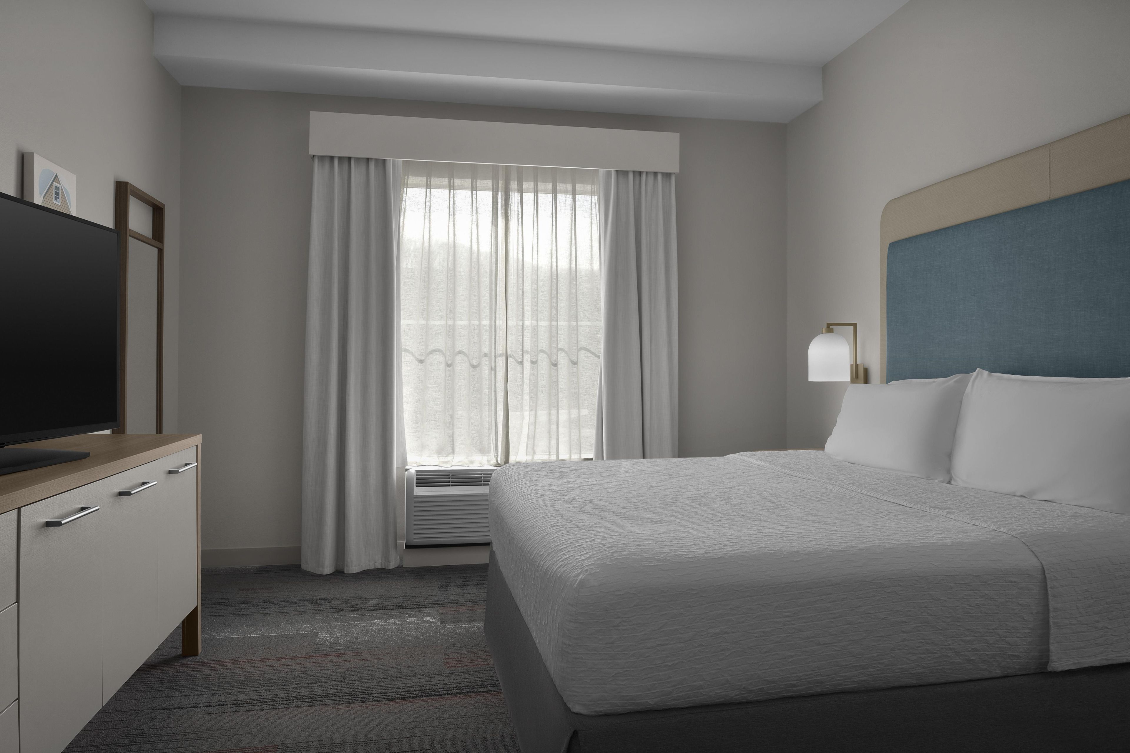 A hotel room with a bed, a TV on a wooden dresser, a window with curtains, and a bedside lamp.