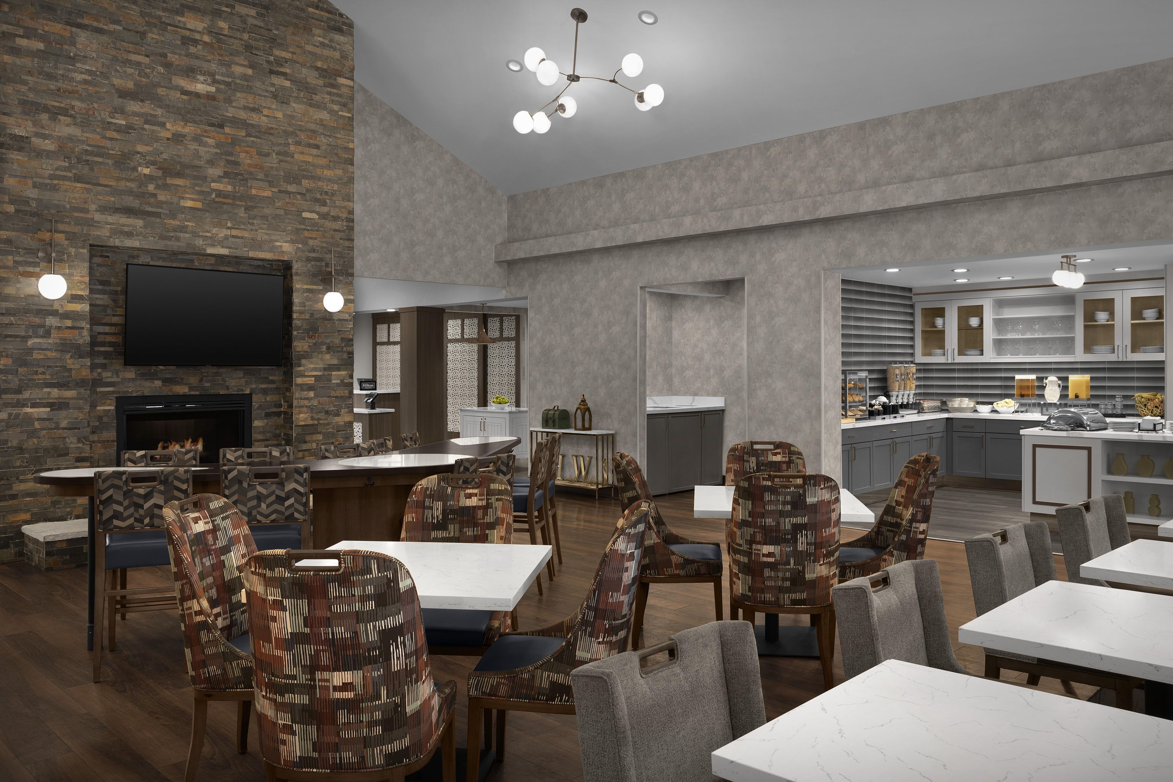 A modern hotel lobby with a stone wall, a flat-screen TV, a fireplace, and a dining area with patterned chairs and tables.