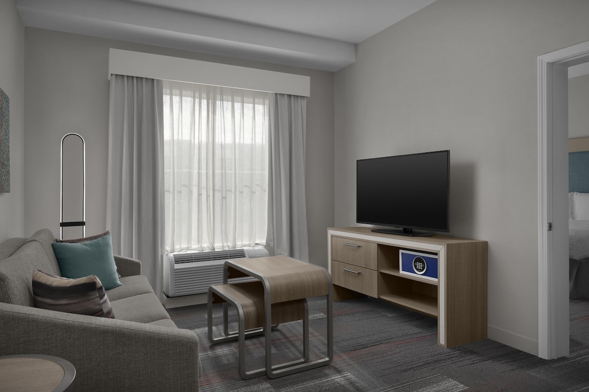 A modern hotel room with a sofa, a TV, a desk, and a large window with curtains.