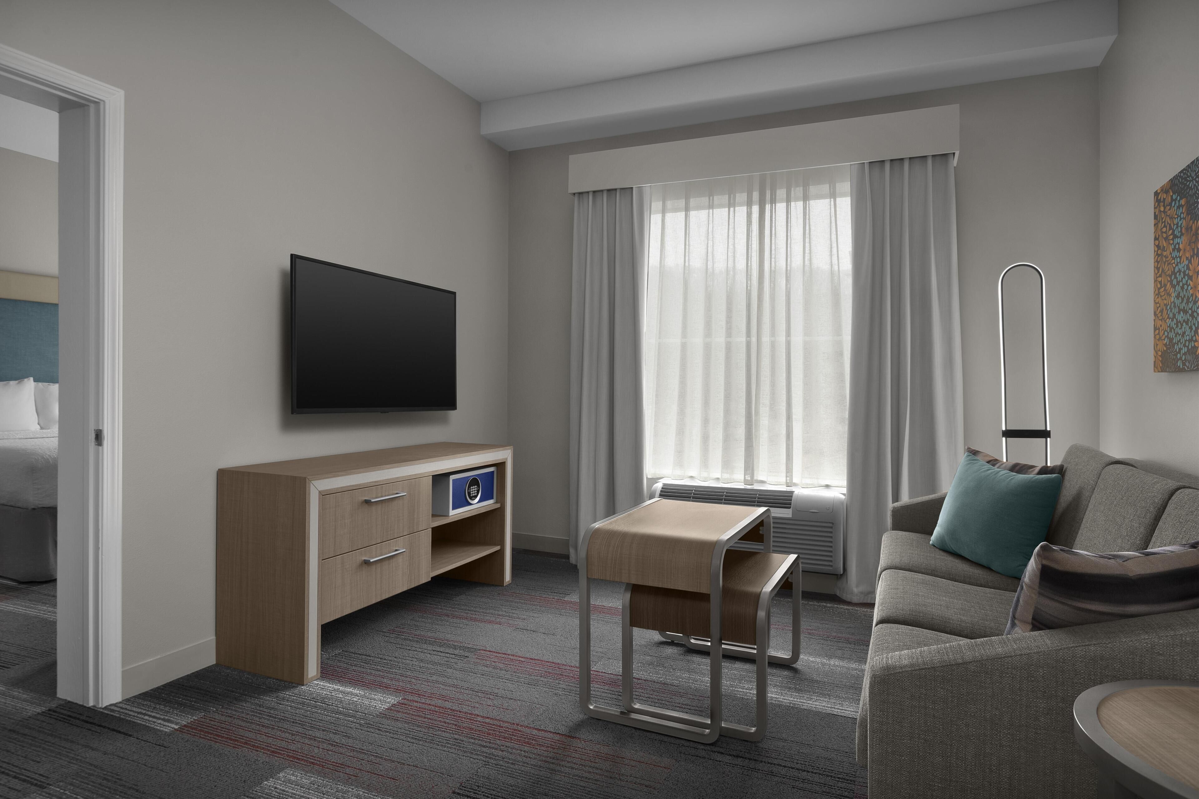 A hotel room with a flat-screen TV, a wooden dresser, a small table, a sofa, and a window with curtains.
