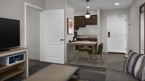 A modern living room with a flat-screen TV, a sofa, a coffee table, and a kitchen area with cabinets and a dining table.