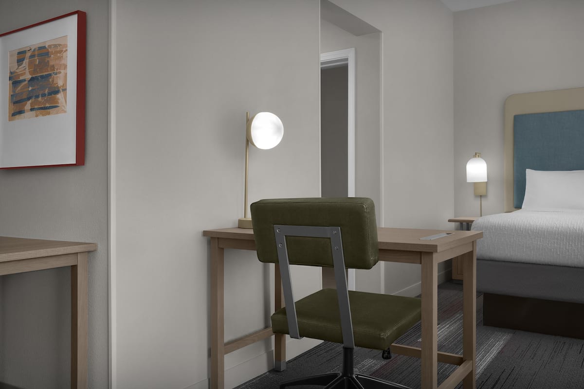 A hotel room with a bed, a desk with a chair, a bedside table with a lamp, and a framed picture on the wall.