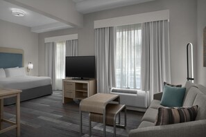 Ein Hotelzimmer mit Bett, Fernseher, Schreibtisch, Sofa und einem Fenster mit Vorhängen.