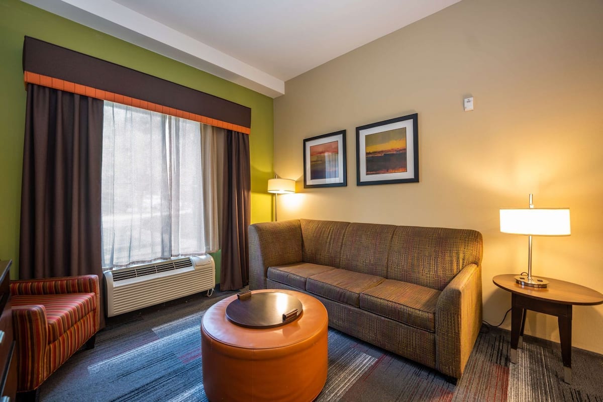 A hotel room with a sofa, a round ottoman, a small table with a lamp, a window with curtains, and two framed pictures on the wall.