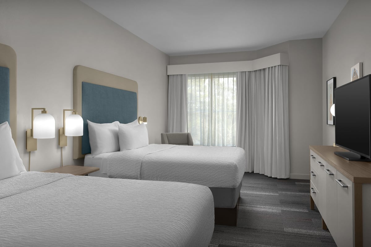 A hotel room with two beds, a TV, a desk, and a window with curtains.