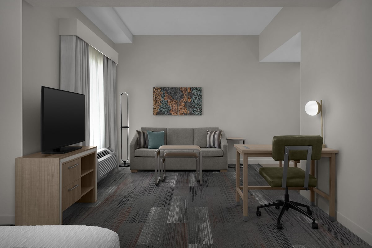 A modern hotel room with a flat-screen TV, a sofa, a coffee table, a desk with a chair, and a painting on the wall.