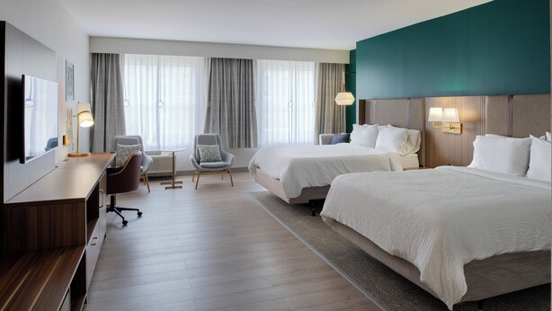 Una habitación de hotel con dos camas, un escritorio, una silla, un televisor y un ventanal con cortinas.
