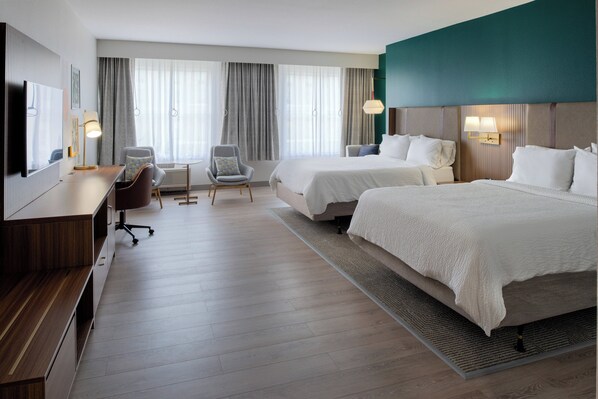 Quarto de hotel com duas camas, uma escrivaninha, uma cadeira, uma televisão e uma janela grande com cortinas.