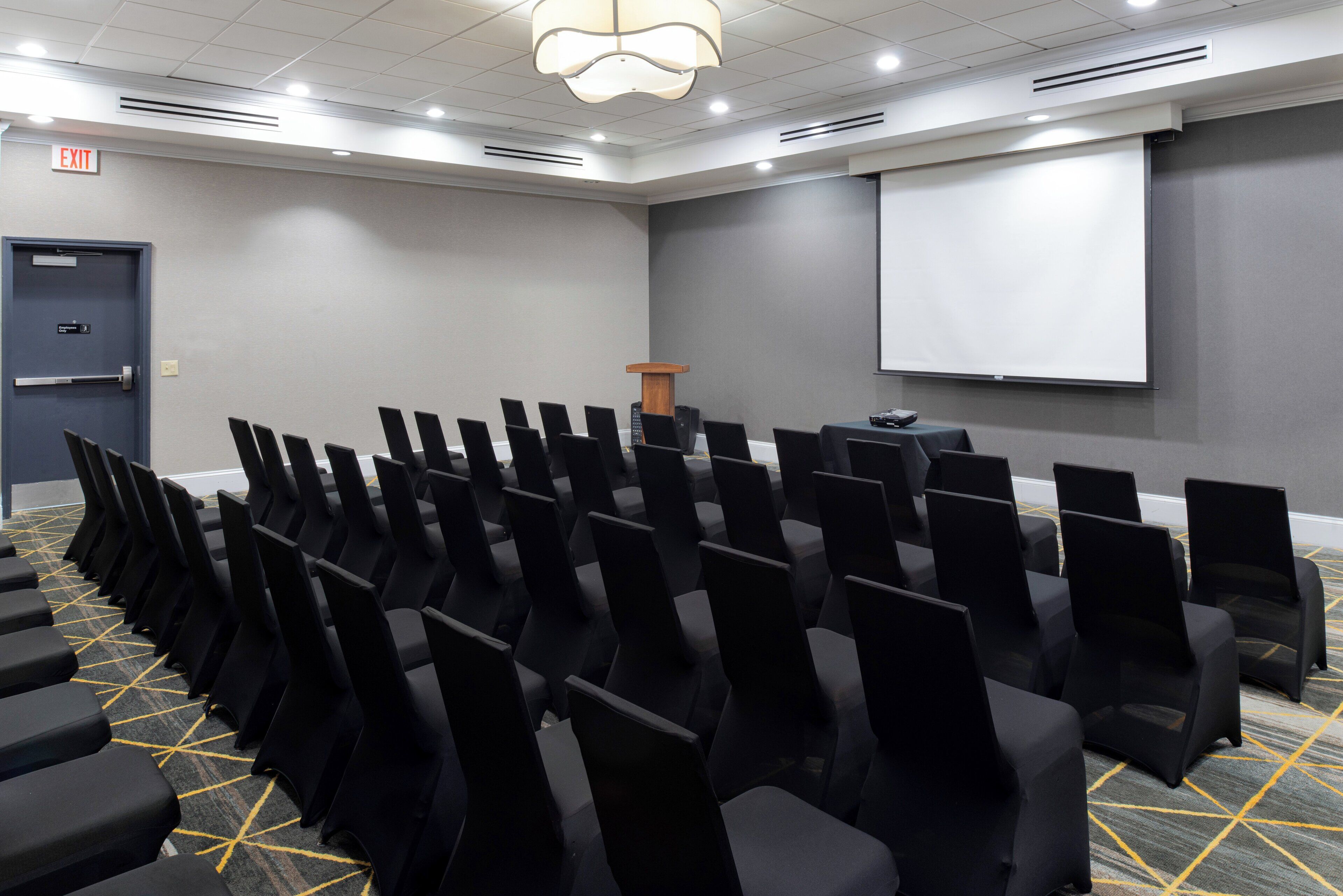 A conference room with rows of black chairs, a podium, a projector screen, and an exit sign.