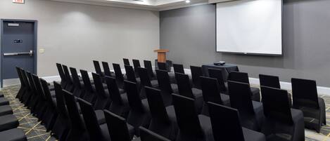 A conference room with rows of black chairs, a podium, a projector screen, and an exit sign.