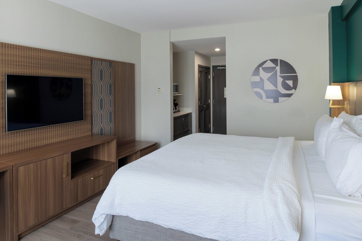 A hotel room with a large bed, a TV mounted on a wooden cabinet, a bathroom visible through an open door, and a wall art piece.