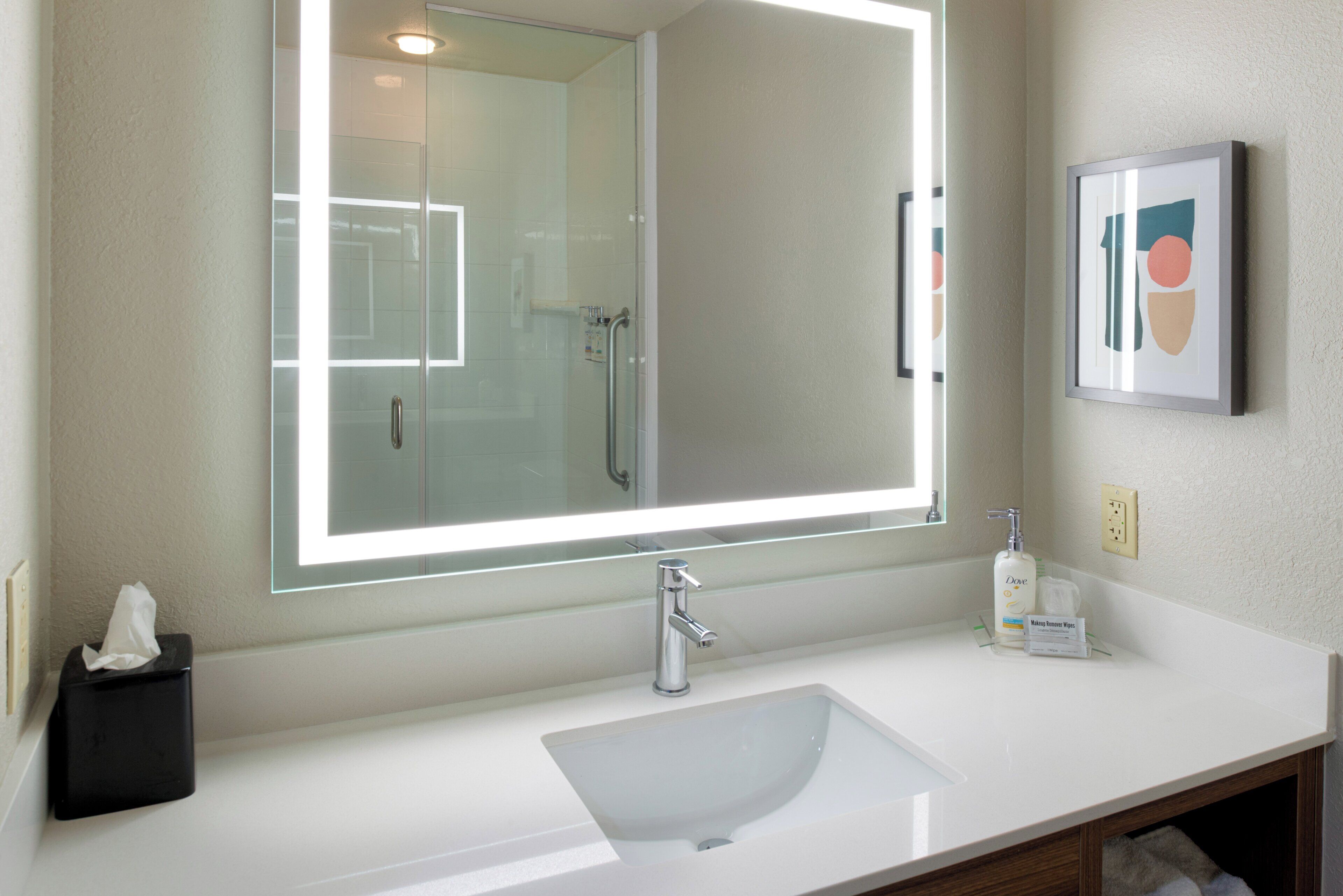 Une salle de bain avec un grand miroir, un lavabo blanc, une douche en verre et une œuvre d’art encadrée au mur.