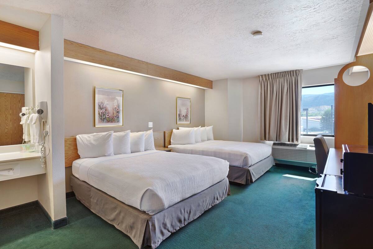 A hotel room with two beds, a desk, a chair, a large window with curtains, and artwork on the walls.