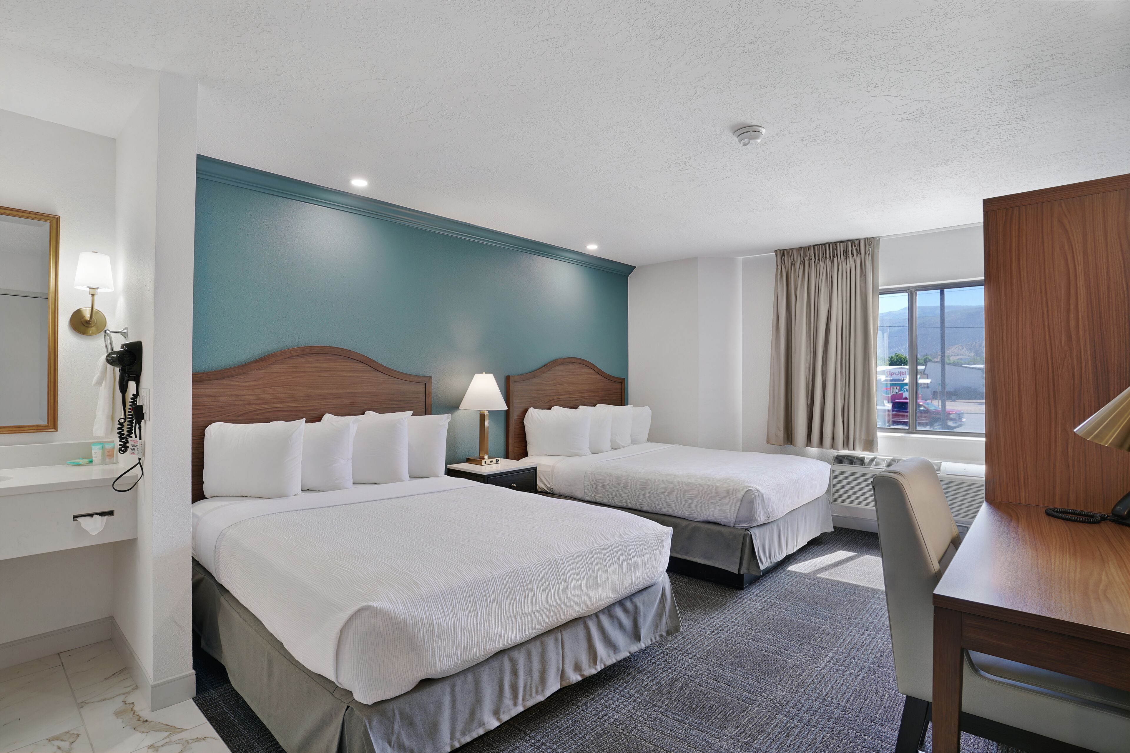 A hotel room with two beds, a desk, a chair, a lamp, a mirror, and a view of the outside.