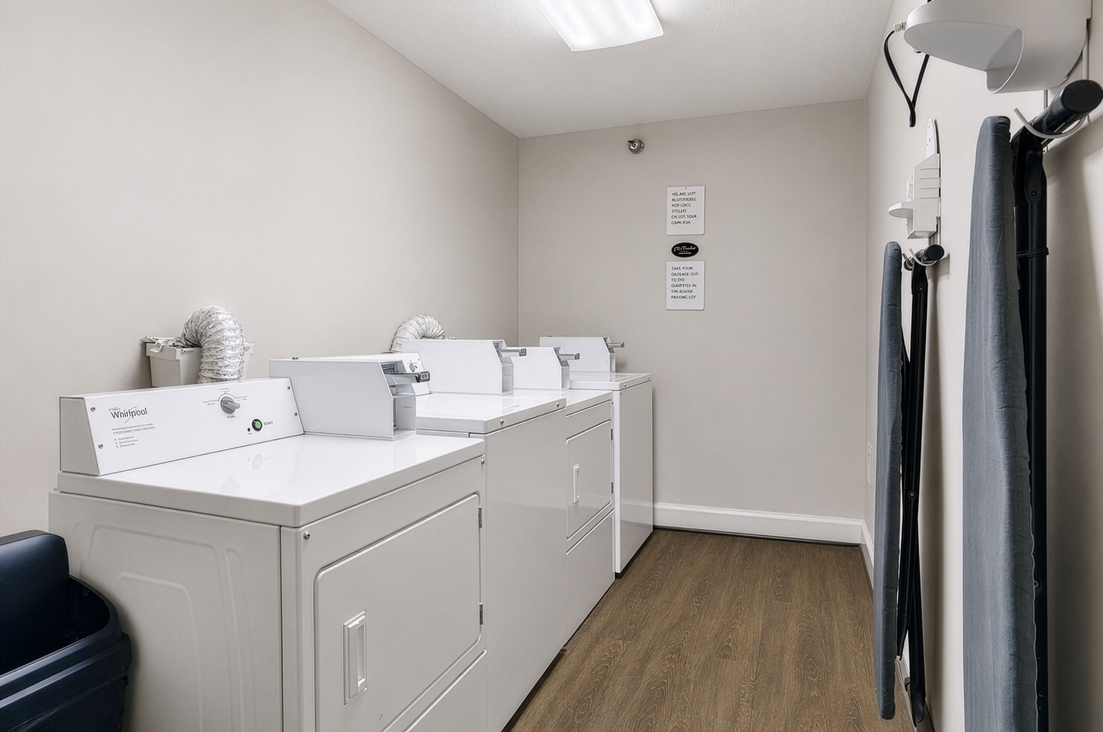 A laundry room with white appliances, a black trash bin, and a wooden floor.