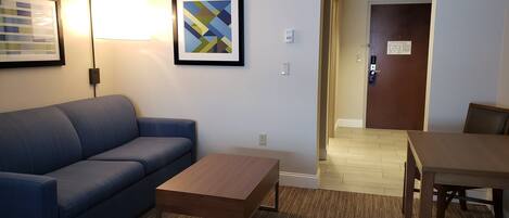 Una habitación de hotel con un sofá azul, una mesa de centro de madera, una lámpara y un cuadro abstracto enmarcado.