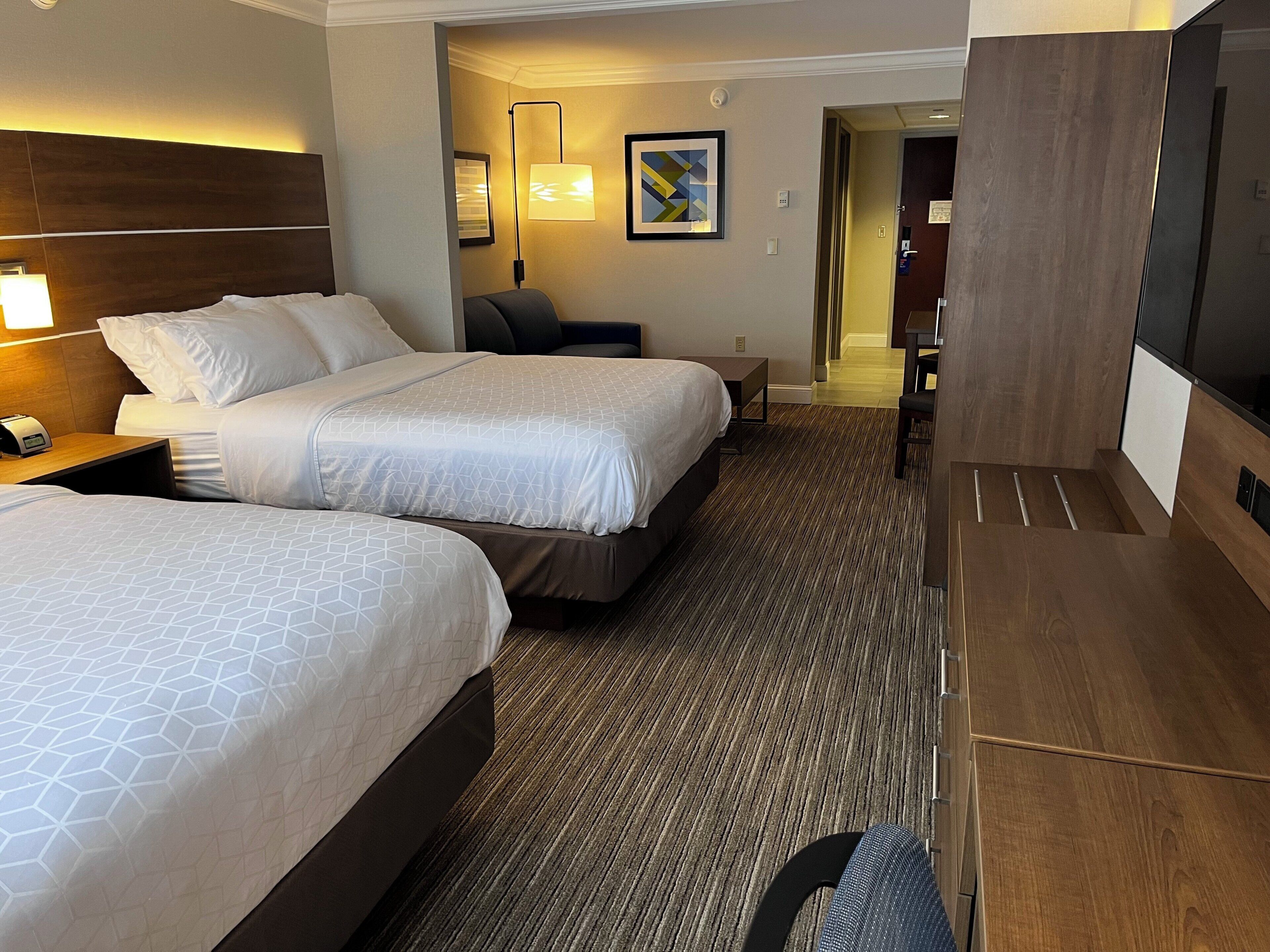 A hotel room with two beds, a desk, a chair, a TV, and a lamp.