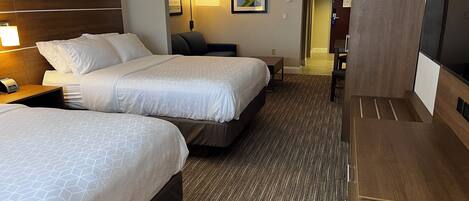 A hotel room with two beds, a desk, a chair, a TV, and a lamp.