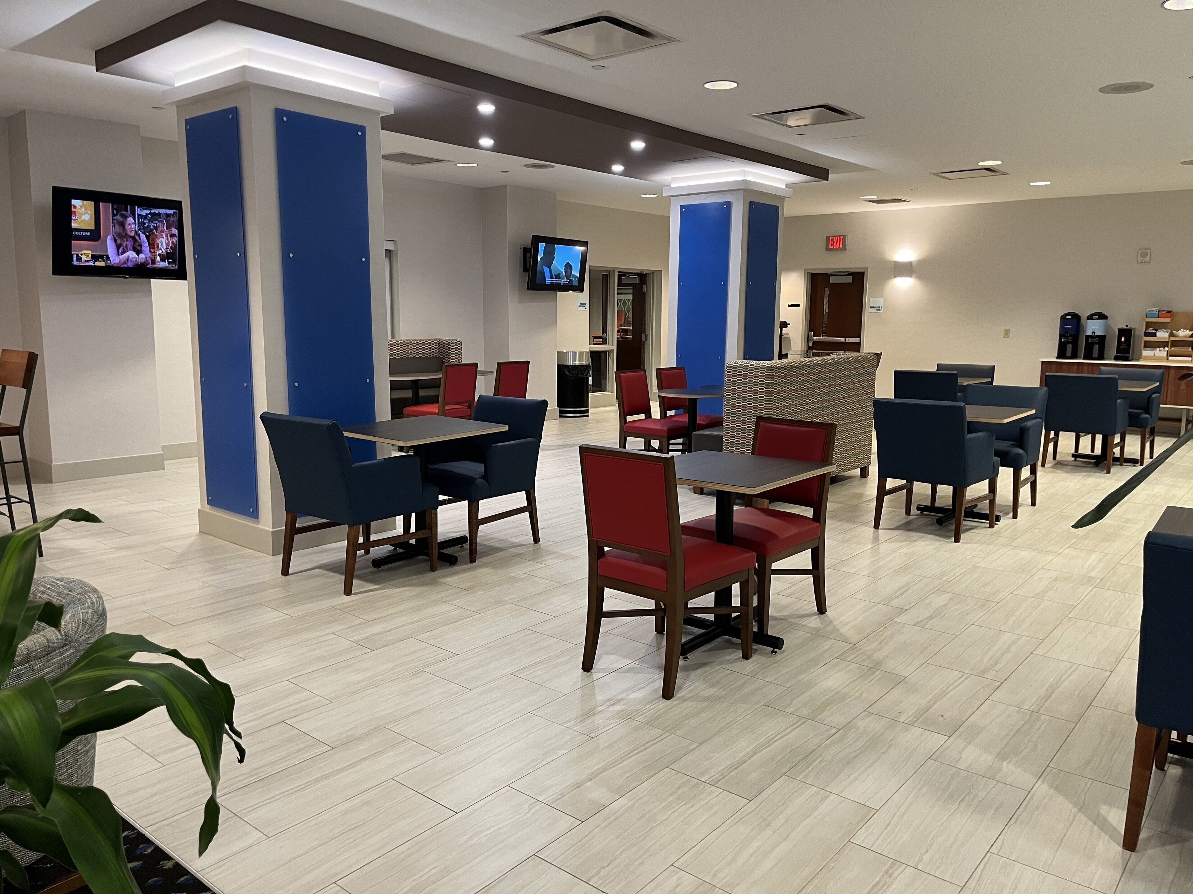 A hotel lobby with multiple seating areas, including tables and chairs, a television mounted on the wall, and a trash can.