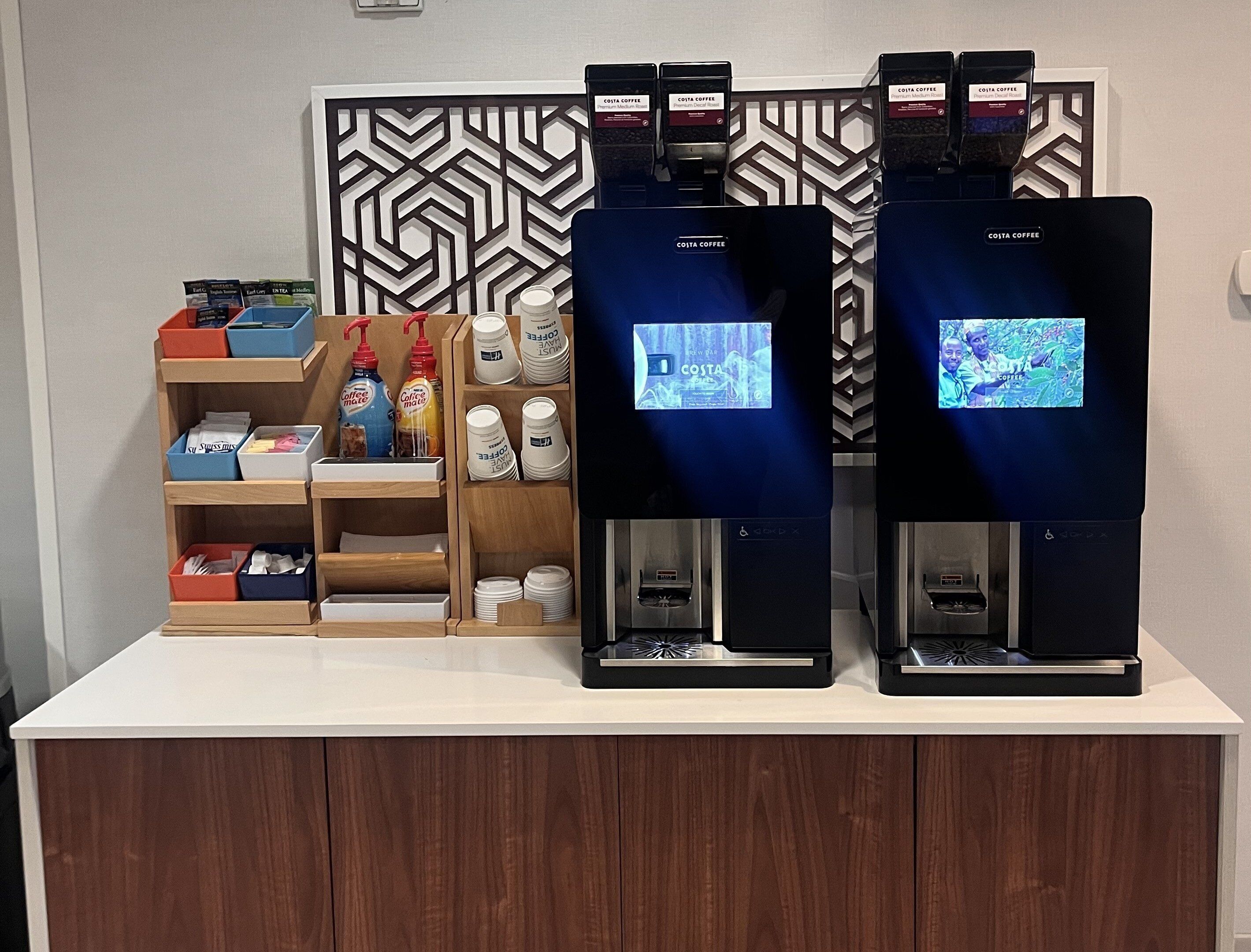 Een Costa Coffee hoek met twee koffiemachines, een presentatie van wegwerpbekers, en een plank met koffiepads en een desinfecterende handgeldispenser.