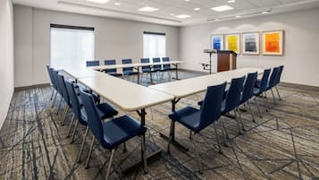 Un salón de conferencias con una larga mesa, sillas azules, un atril y obras enmarcadas en la pared.