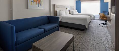 Habitación de hotel con sofá azul, una mesita de madera, dos camas, un escritorio y un televisor.