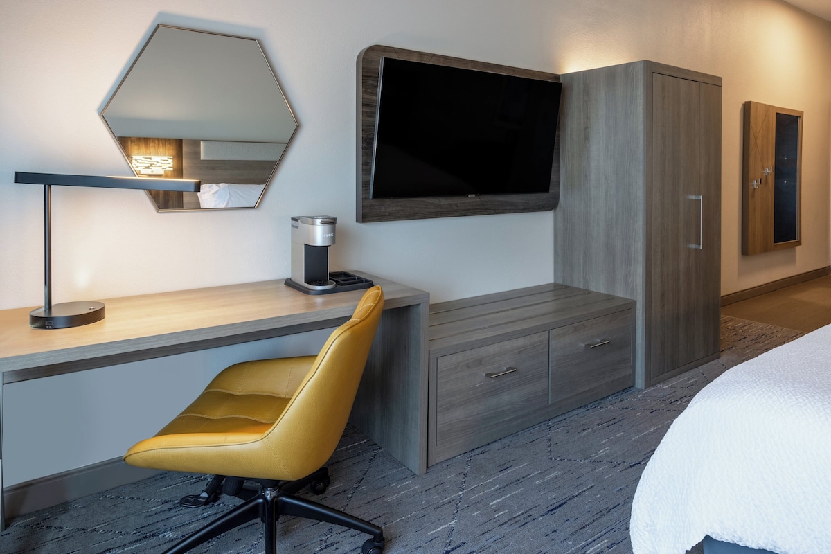 A modern hotel room with a large bed, a desk with a chair, a flat-screen TV, a wardrobe, and a mirror.