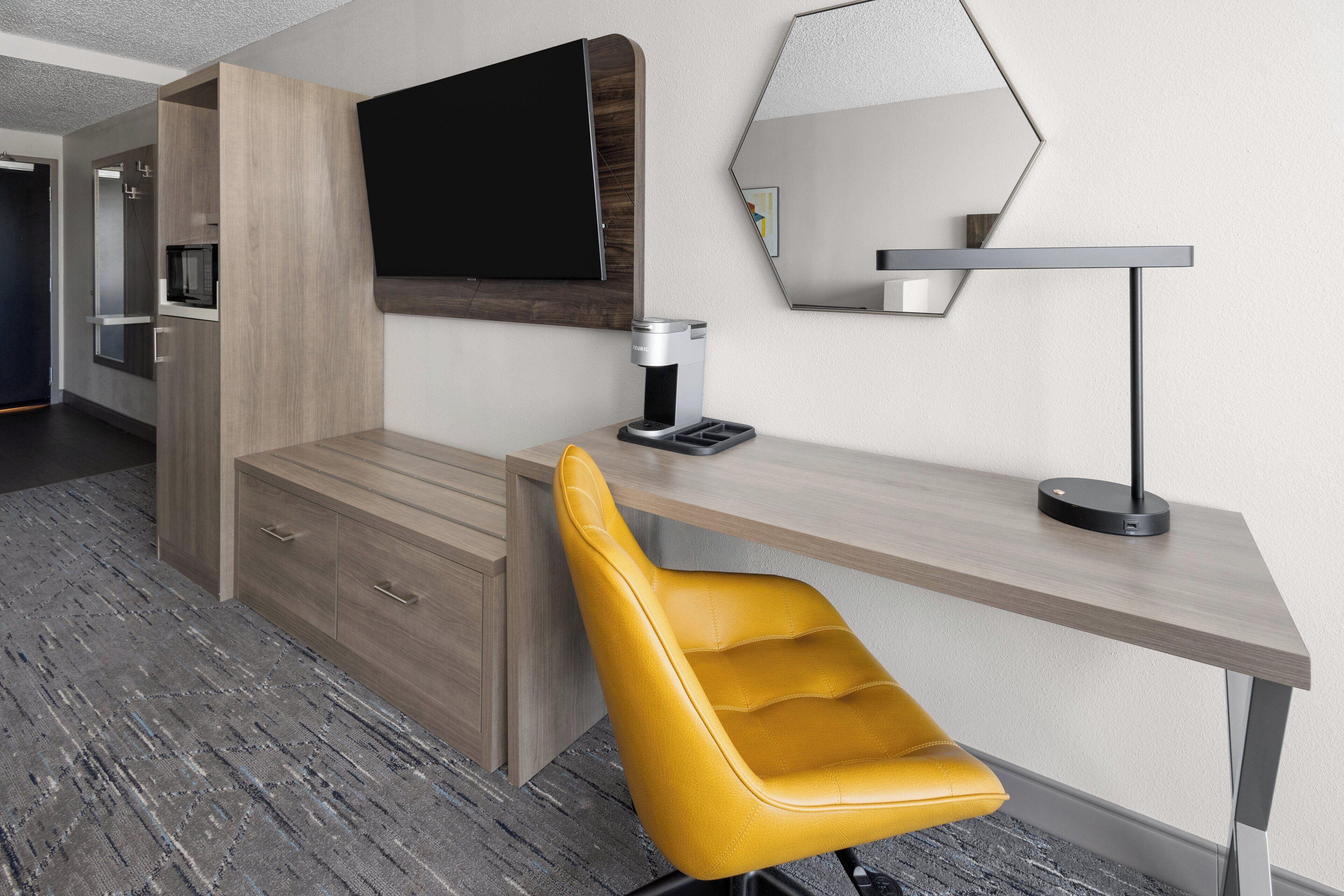 Una habitación moderna con un escritorio de madera, una silla amarilla, un televisor de pantalla plana, una cafetera y un espejo.