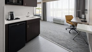 A modern hotel room with a kitchenette, including a microwave, coffee maker, and refrigerator. There is a dining area with a table and a chair, and a large window providing a view of the outdoors.