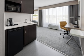 A modern hotel room with a kitchenette, including a microwave, coffee maker, and refrigerator. There is a dining area with a table and a chair, and a large window providing a view of the outdoors.