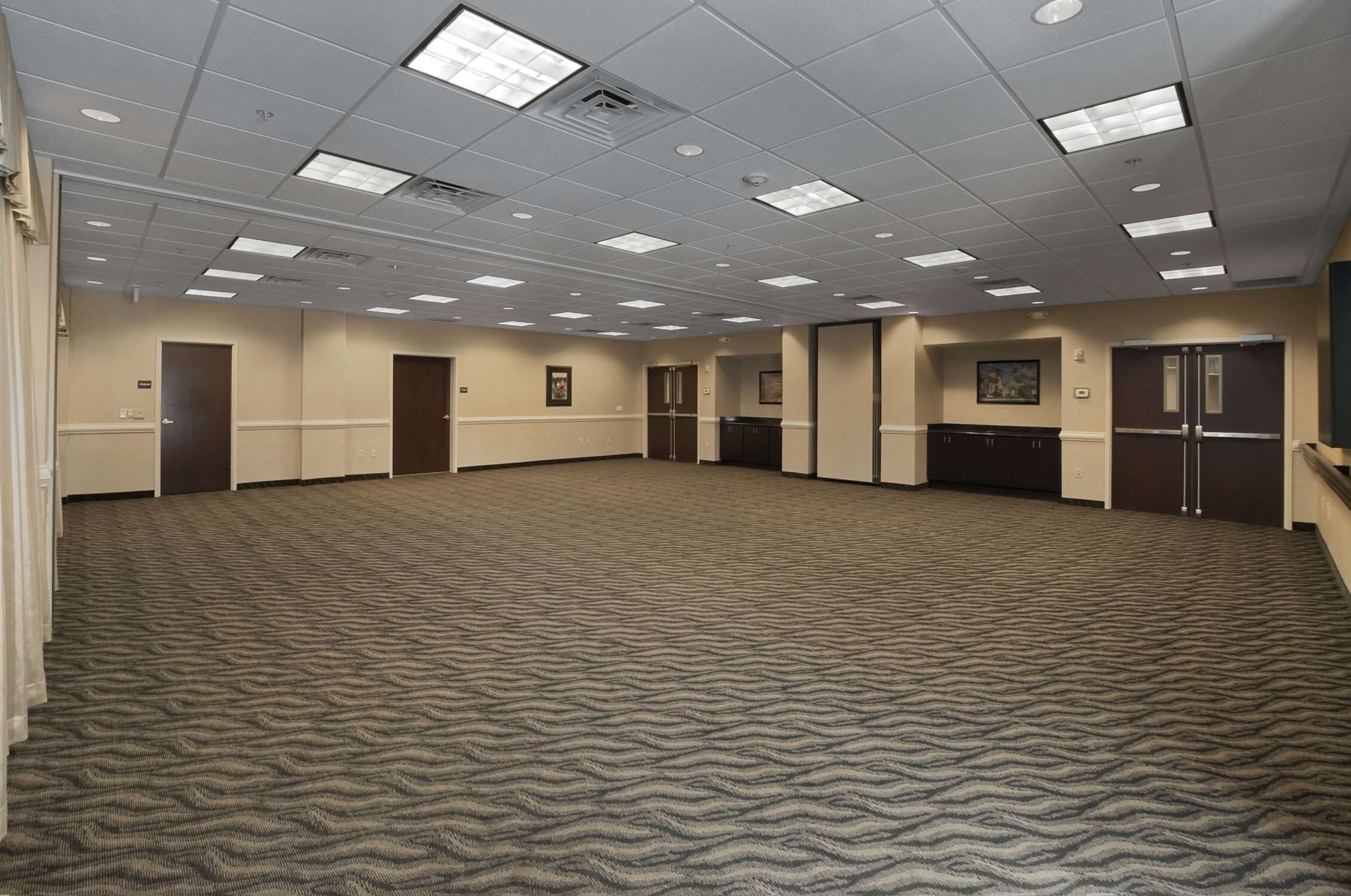 Un amplio salón de conferencias con alfombra, techo alto, iluminación empotrada y una distribución simétrica, con puertas y una zona de kitchenette.