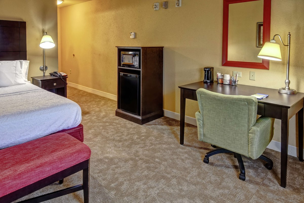 A hotel room with a bed, a desk, a chair, a minibar, and a lamp.