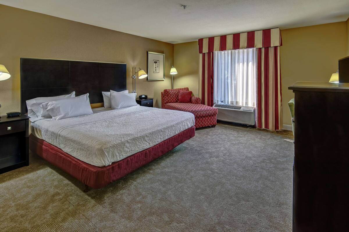 A hotel room with a large bed, a bedside table, a chair, a desk, and a window with curtains.