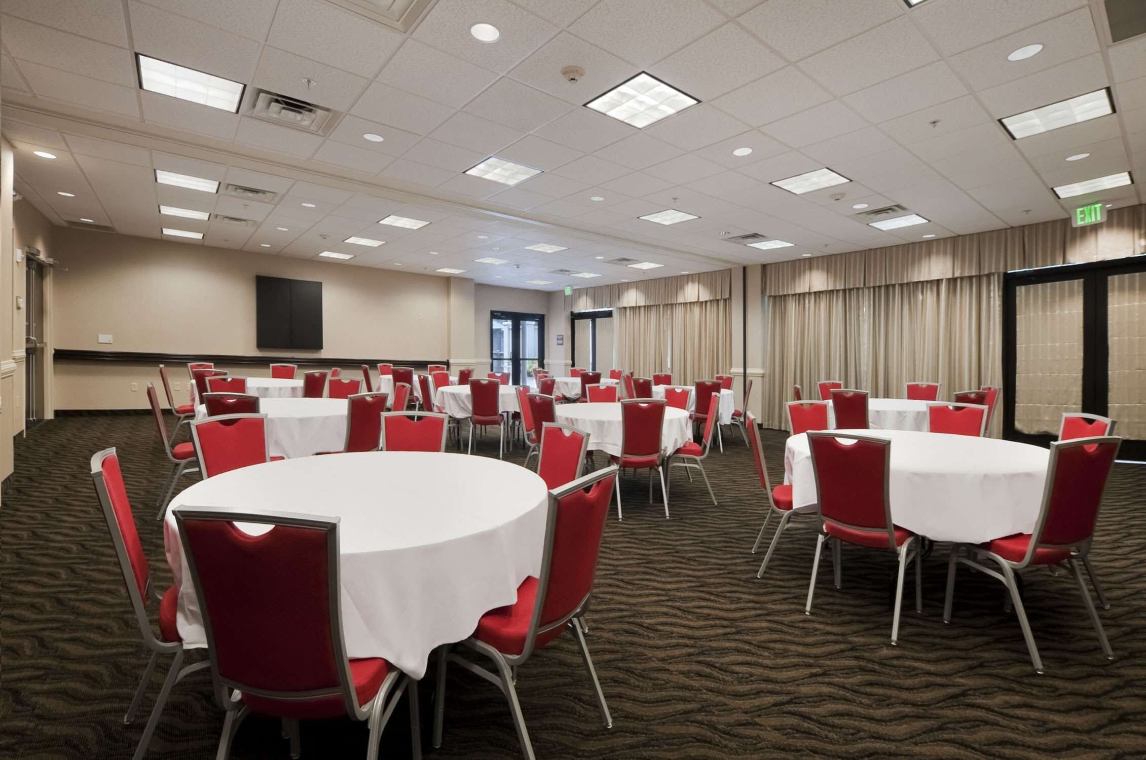 A conference room with multiple round tables and red chairs, carpeted floor, and a large screen on the wall.