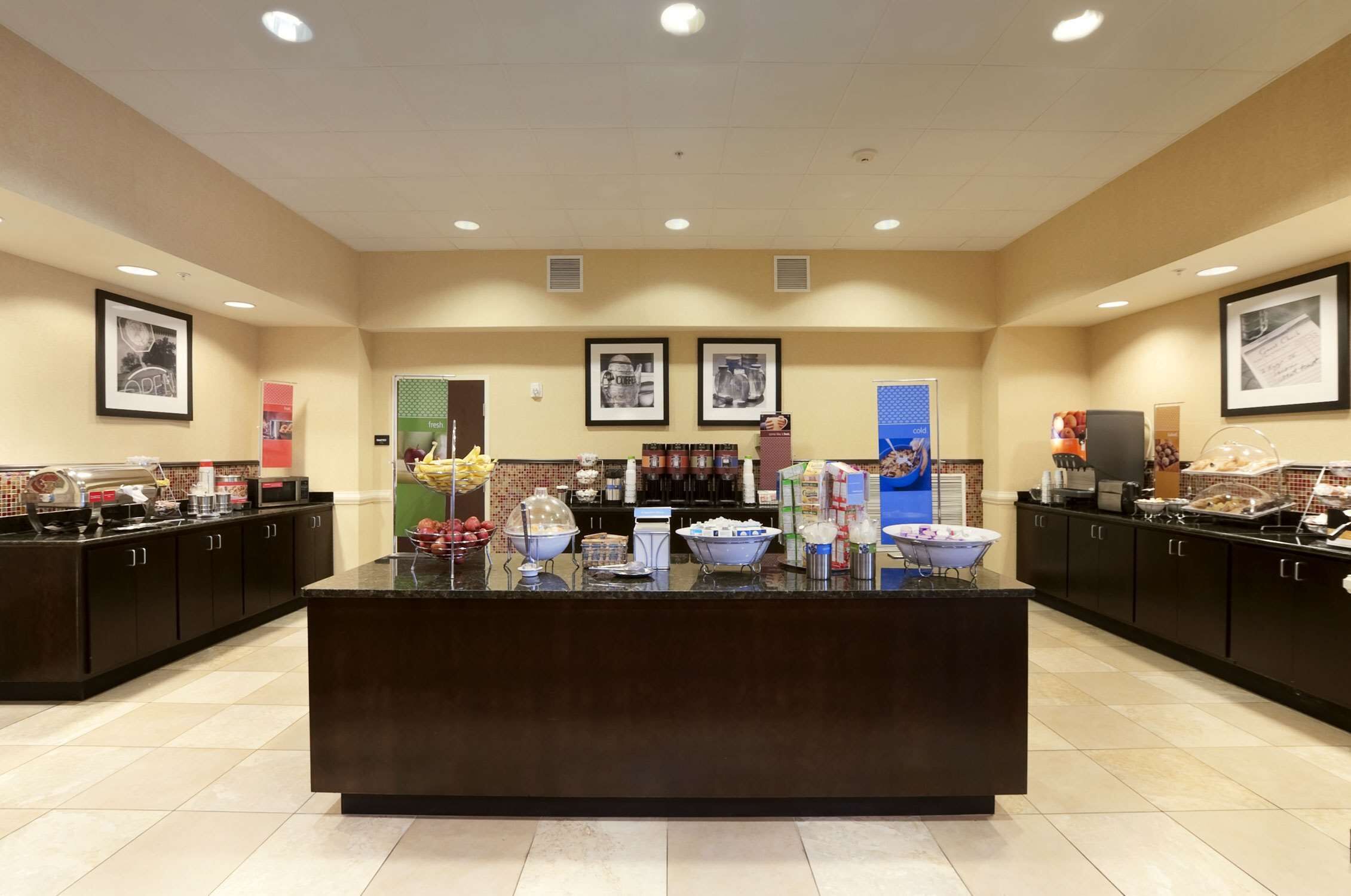 A spacious breakfast area with a central counter, various food stations, and framed pictures on the walls.